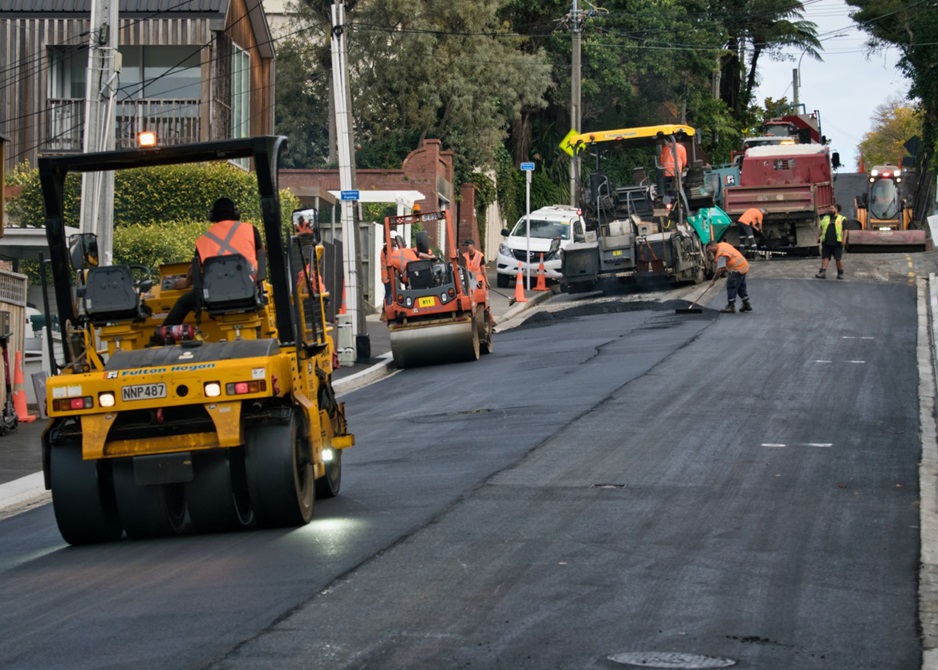Workers and machinery surfacing a road with asphalt. A steam roller is smoothing the surface.