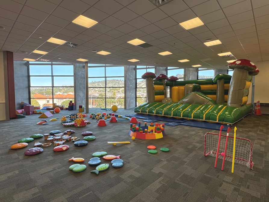 A bouncy castle surrounded by soft obstacles and colourful toys for small children, set up inside a room.