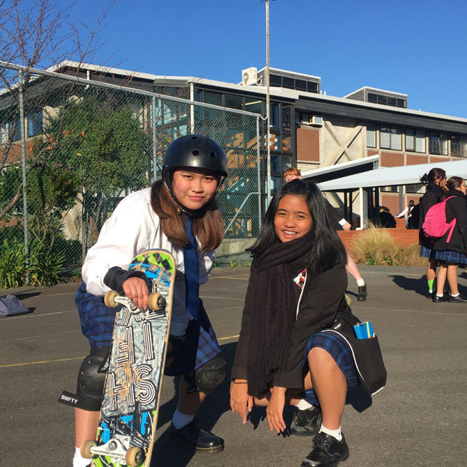 Two young women posing happily with their skateboards at school.