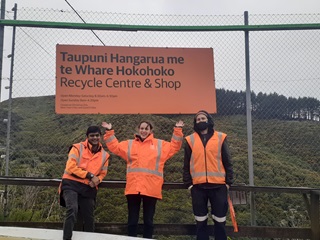 Three smiling staff members standing in front of the fence and ‘Taupuni Hangarua me te Whare Hokohoko Recycle Centre and Shop’ sign at Ōwhiro Bay. 