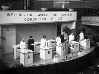 Wellington apple pie contest conducted by radio station 2ZB in the Town Hall, Wellington, on 23 April 1940. Radio personality Aunt Daisy (Maud Basham) stands at the front of the stage.