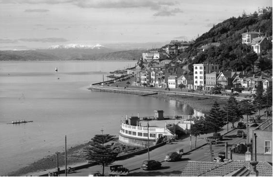 A black and white photo of Oriental Parade shot from behind. It shows the Band Rotunda and a Waka in the harbour.