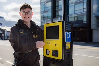 A Council parking officer standing next to a yellow and black Pay by Plate parking meter on a street in Wellington.