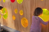 Children looking through colourful perspex 'windows' in plywood hoarding.