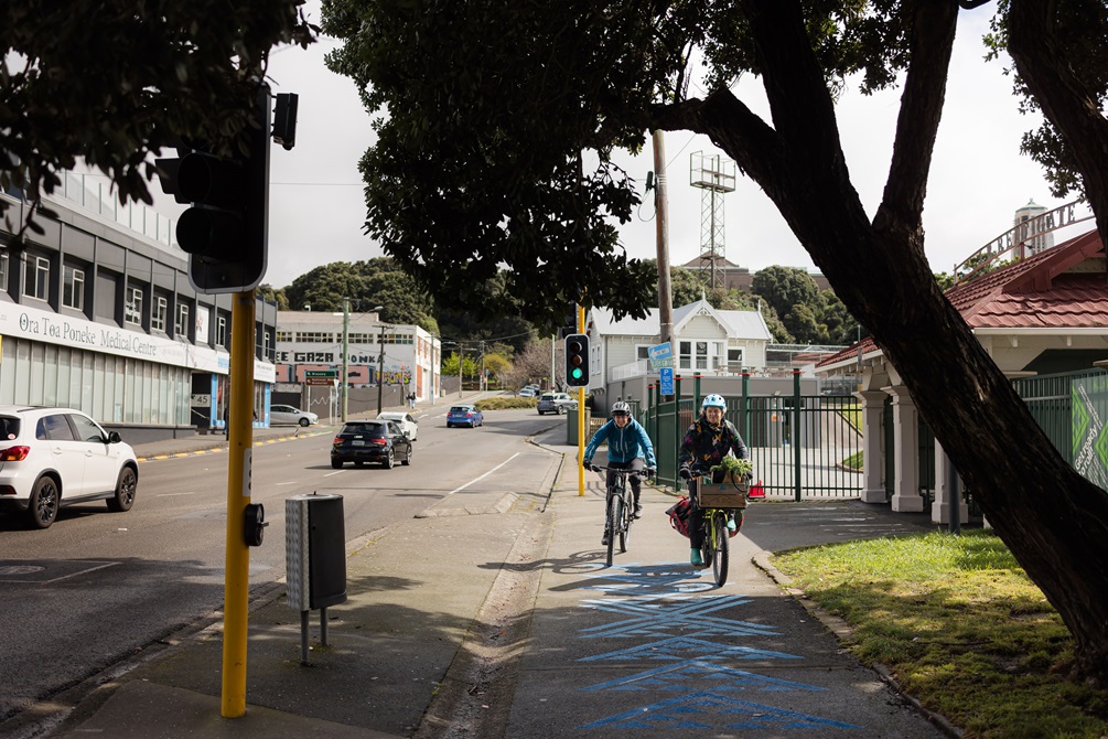 Two people on bikes on the Southern side of the Basin reserve, blue markings can be seen on the path they are riding on.