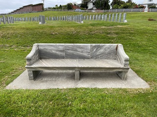 A bench made of marble in the foreground, with gravestones in the background.