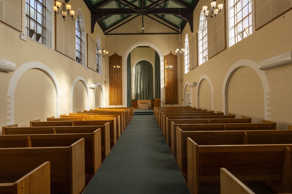 Entrance of main chapel, showing rows of wooden seats, and large windows with sun coming in from the right.