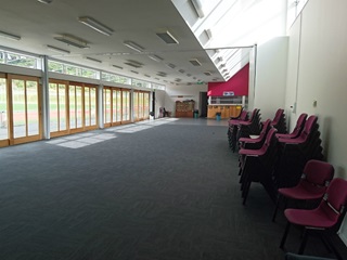 An empty community hall with chairs stacked on the right-hand side and windows down the left-hand side.