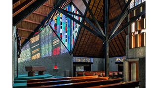 Photo of the interior of Futuna church featuring wooden pews, a high ceiling made of wooden beams and planks, and large coloured-glass panels.