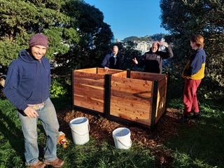 People standing around two big wooden compost bins, with white plastic buckets in the foreground.