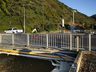 Photo taken on the seaward side of the bridge over the Kaiwharawhara Stream on Hutt Road showing the stream underneath, the bridge handrail, and beyond that, someone biking over the bridge and a white ute travelling along the road..