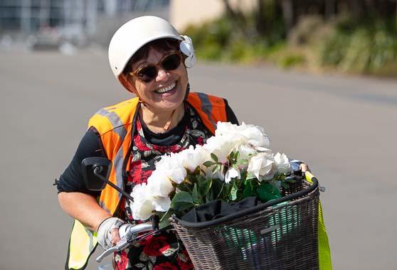 Woman on a bicycle, wearing a helmet and a high-visibility vest, carrying a bunch of white flowers on the front basket.