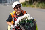 Woman on a bicycle, wearing a helmet and a high-visibility vest, carrying a bunch of white flowers on the front basket.