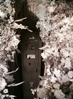 Black and white photograph of a coffin being lowered into a grave surrounded by flowers. 