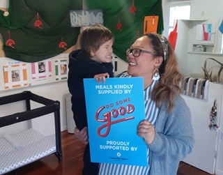 Hilda Sue, Head Teacher from Ngahauranga Kohanga Reo, holding toddler Henare and a big blue poster promoting 'Do Some Good', a free lunch programme for pupils which has been rolled out at the school. The pair are smiling at each other.