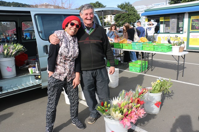 Tulelei and Tom Hayes, in an embrace smiling at the camera, in front of their van, with buckets of fresh flowers and citrus fruit surrounding them at Harbourside Market. 