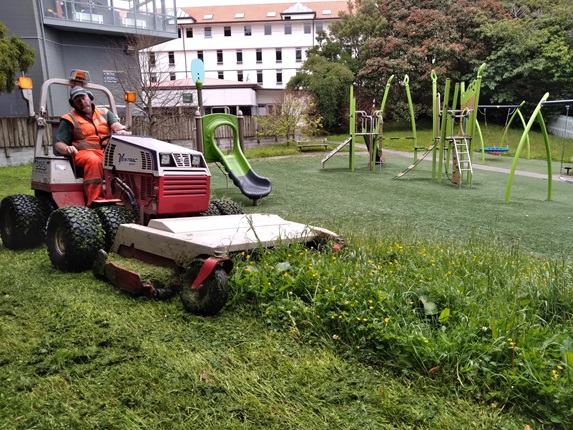 A Council mowing expert giving a community park some TLC on a ride-on lawn mower. Playground equipment and flats can be seen in background.