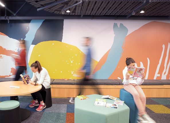 Two children sitting at separate tables, each engrossed in a book, with the bright Te Awe Library mural behind them and blurred people walking by.
