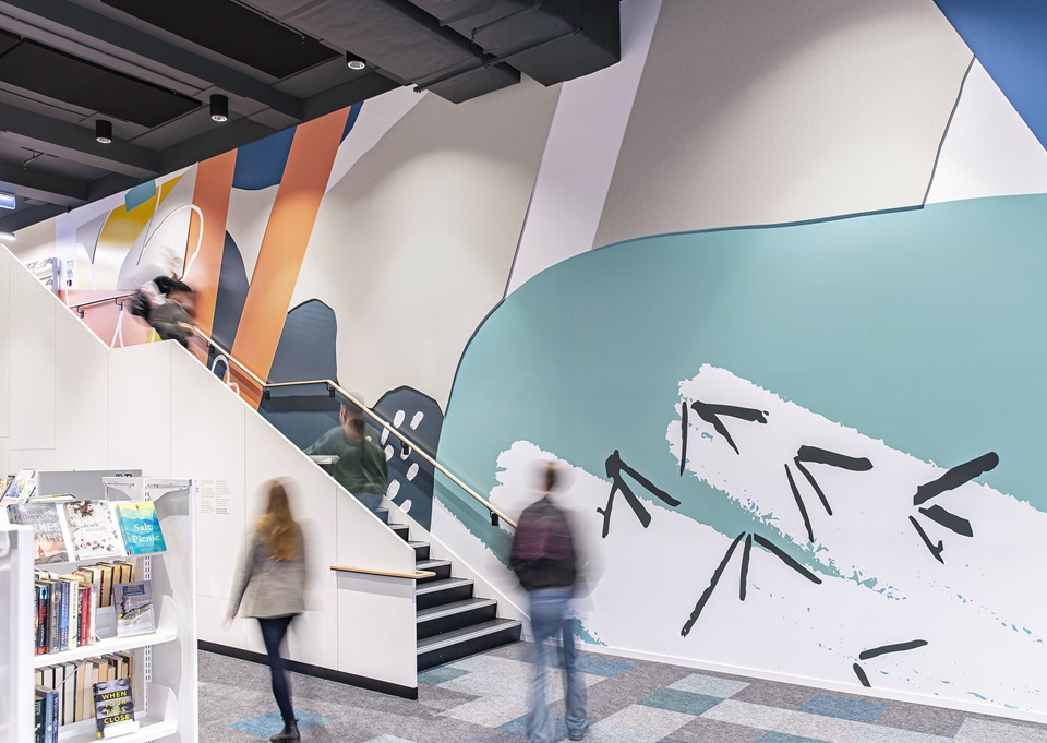 A floor-to-ceiling mural, featuring bright colours and nature-inspired shapes like bird footprints, alongside the stairwell at Te Awe Library.