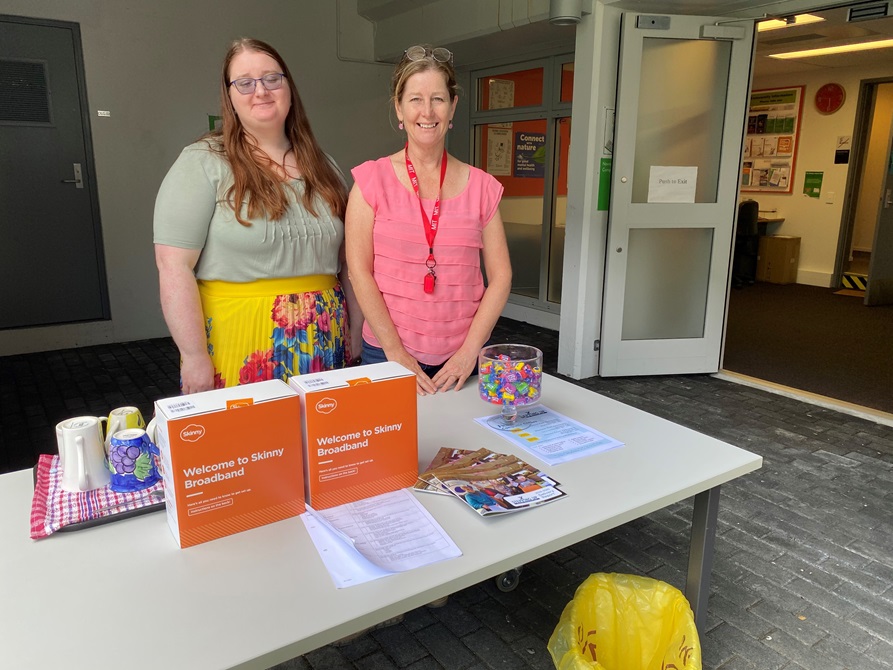 Two smiling women in lovely bright clothing standing at a table with information about Skinny Broadband and lollies on offer.