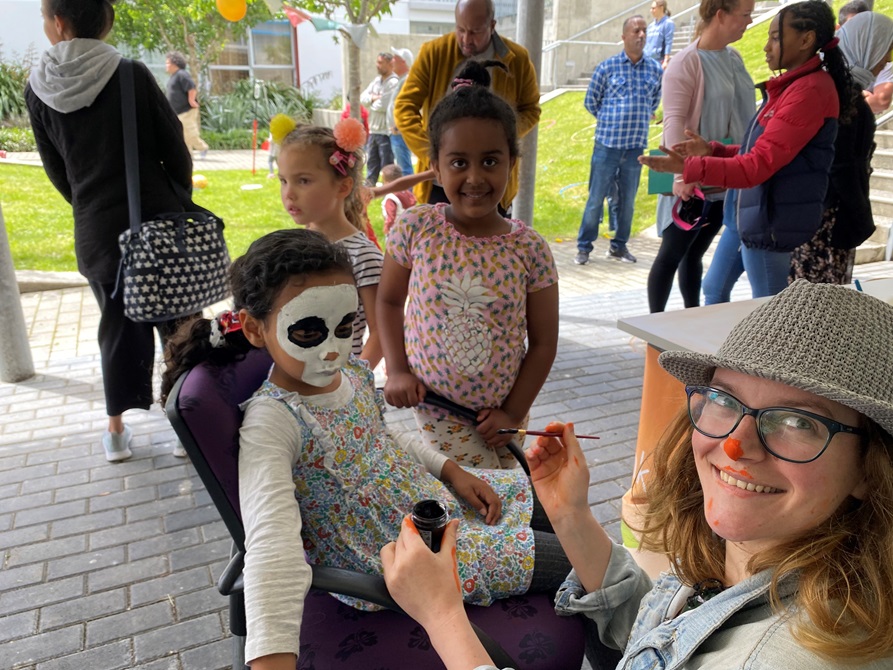 Three children eagerly awaiting their turn with the face painter, who has paintbrush in hand and is smiling at camera.