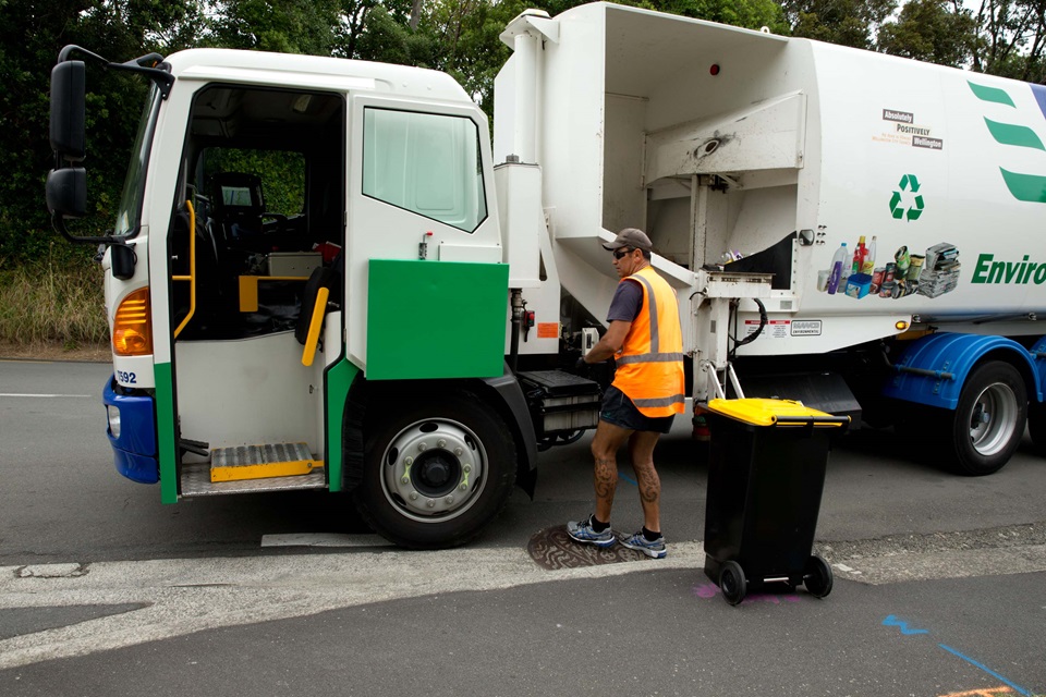 A recycling truck with a male staff member in a high vis, cap and sunnies, standing beside a wheelie bin on a dry road.