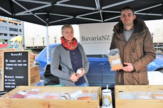 Young couple Lena Donandt and Sebastian Nebel standing on either side of their business sign, BavariaNZ, with their products, handmade German-style sausages, laid out in front of them in their marquee stall.