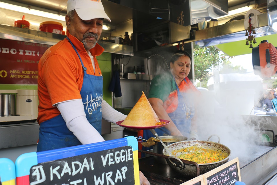 Balu Rajagopal wearing bright orange and blue serving up South Indian dosa and curry onto a plate, with his wife Shree Balasubramaniam to his right spreading batter onto a hotplate which is obscured by steam. 
