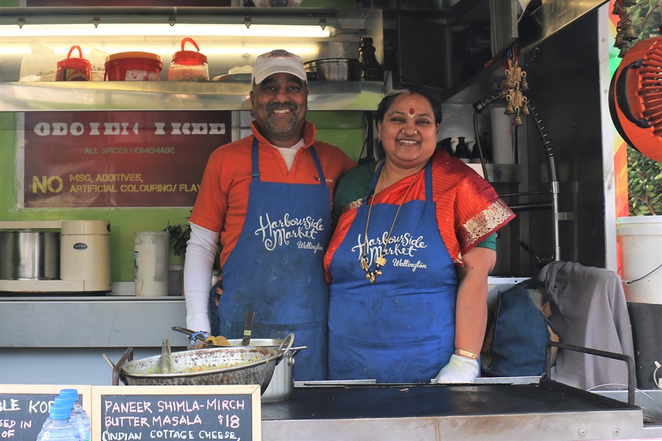 Husband and wife Balu Rajagopal and Shree Balasubramaniam, in orange t-shits and blue aprons, smiling from their food caravan, where they serve South Indian vegetarian food.