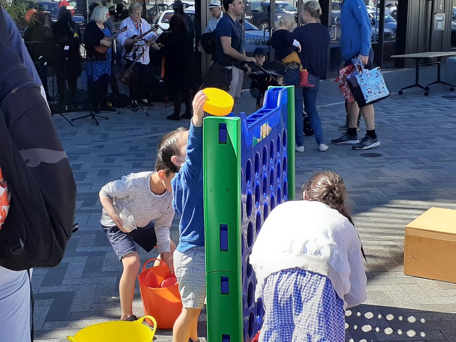 Three children enjoy a game of giant Connect Four, with a band playing music in the background, standing in front of Waitohi.