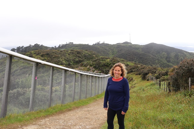 Brooklyn resident Mel Beirne in her running gear, standing to the right of the Zealandia fence line on the Brooklyn Wind Turbine Route, with hills and the turbine behind her.