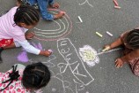 An overhead shot of children with braids and ponytails drawing shapes and rainbows in colourful chalk on the concrete. 