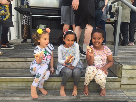 Three children named Amelia, 5, Libby, 6, and Makeda, 5, sitting in a row on wooden steps holding sausage sizzles with adults and a barbecue behind them. 