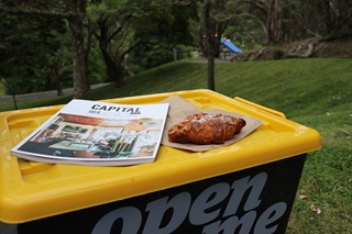 A large green plastic container with a Capital magazine and a croissant sitting on top of the container's yellow lid, placed in a grassy area with a children's playground in background.