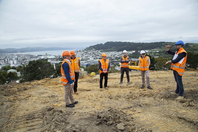 Six men in high-vis vests standing on top of the hillside above Prince of Wales Park, where construction has begun on Wellington's new Omāroro Reservoir, with city and harbour in background.