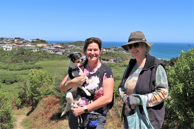 Annie Yeates, holding her Fox Terrier, and Jenny Hartley standing in the sun on the top of Tawatawa Bush Track in Owhiro Bay, with housing and the ocean in the background. 