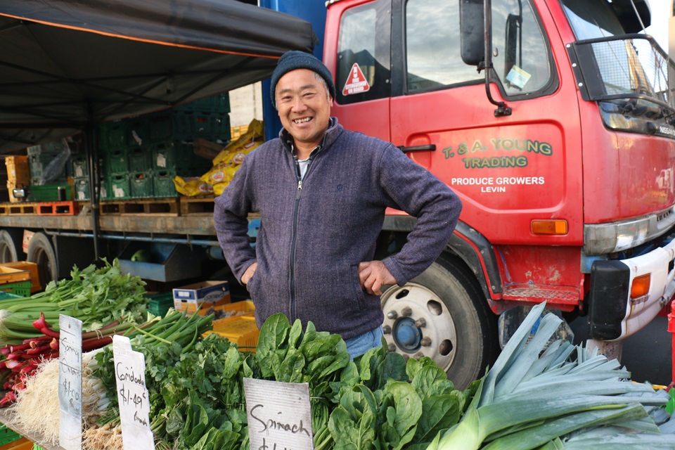 Tommy Young smiling with his hands on his hips, standing in front of his red truck, with a table of his home-grown vegetables in front of him at the Harbourside Market.
