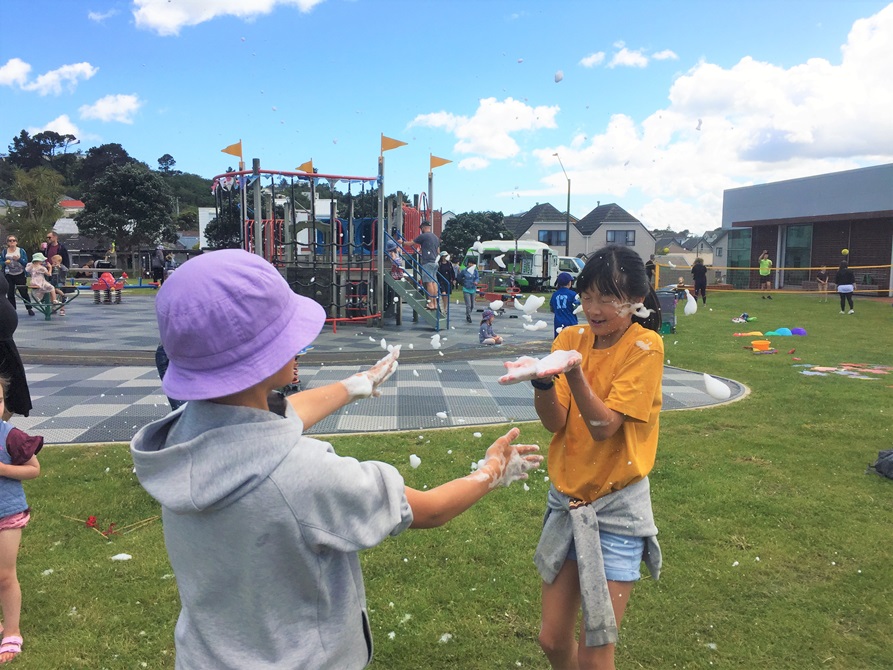 A child in a purple hat, and a girl in a yellow t-shirt, having fun with bubbles in front of the playground at Waitohi.