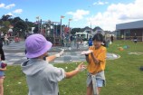 A child in a purple hat, and a girl in a yellow t-shirt, having fun with bubbles in front of the playground at Waitohi.