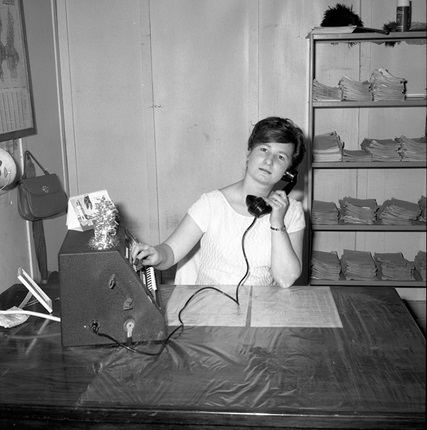 Black and white photo of woman using old telephone at desk in an office. 