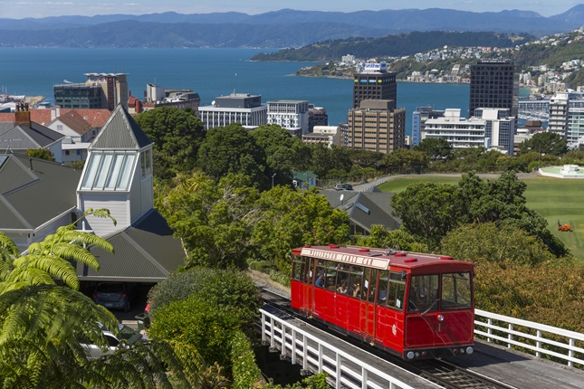 A shot of the red Cable Car on a sunny day, with Wellington buildings and the harbour in the background.