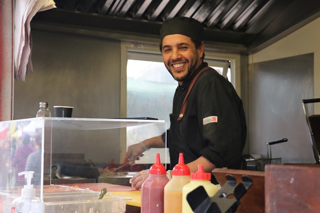 Hassan El Kour, dressed in a black cheffing uniform, smiling while he cooks up traditional Moroccan cuisine from his food truck.