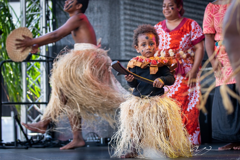A young boy in tradition dress on stage at Wellington Pasifika Festival with others dancing around him.