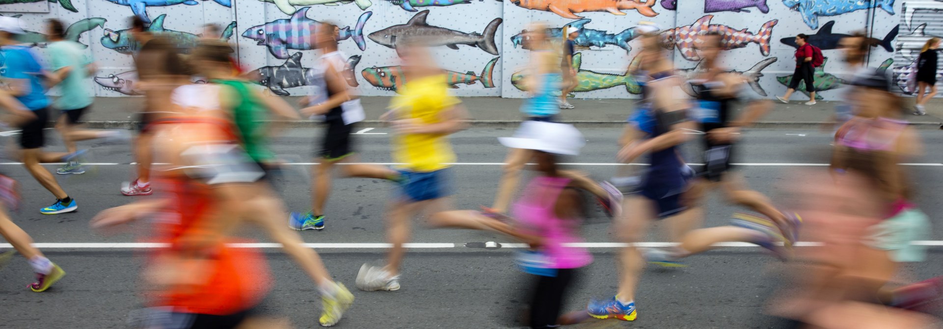Blurred runners in bright activewear, jogging past the colourful shark mural opposite Waitangi Park.