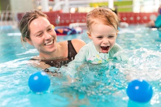 A woman in a swimsuit holding her young child as they smile and play with two blue balls in the pool.