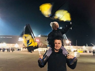 A young boy on his father's shoulders, both waving Hurricanes flags in support of the Wellington rugby team, outside Sky Stadium at night.