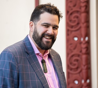 Karepa Wall, in a pink shirt and a smart blue suit, wearing his ponamu, and smiling, pictured from the chest up in front of the engraved side of a marae.