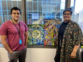 Wellington City Council Contact Centre staff members, Matai and Enisha, standing on either side of two colourful paintings with glass and high rises in the background.