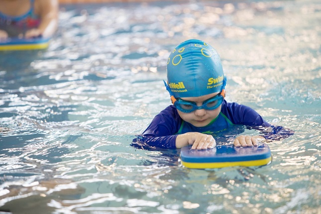 A young child with blue swimsuit, swimming cap and googles on, concentrating while using a flutter board in the pool.