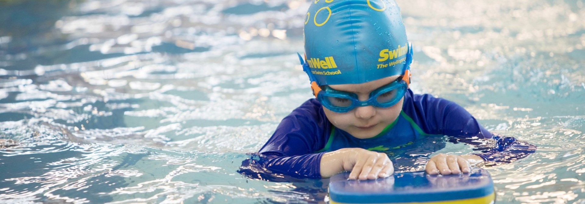 A young child with blue swimsuit, swimming cap and googles on, concentrating while using a flutter board in the pool.